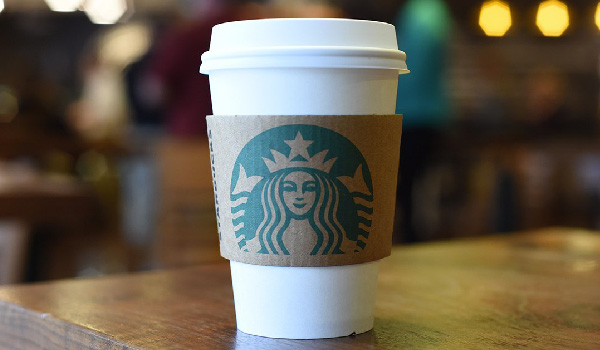 a photo of a Starbucks coffee in a papercup with a brown protective cardboard showing the logo of Starbucks, standing on a wooden table with a blurred background of lights. Beautiful.